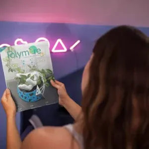 Femme de dos tenant le magazine Polymère & Co N°37 devant elle. Derrière le magazine il y a un mur avec des néons roses de formes géométriques