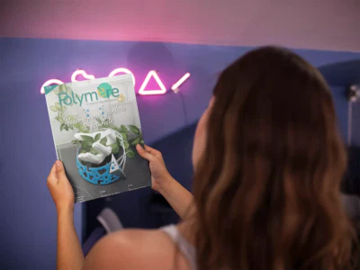 Femme de dos tenant le magazine Polymère & Co N°37 devant elle. Derrière le magazine il y a un mur avec des néons roses de formes géométriques